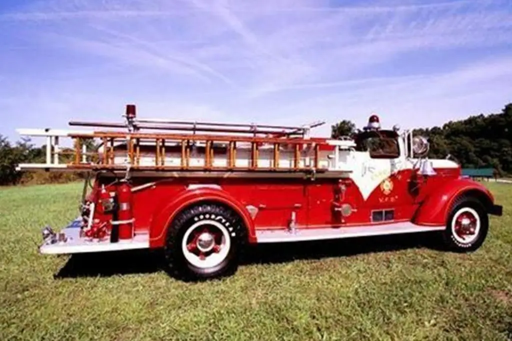 A vintage red fire truck with a wooden ladder and various equipment mounted on top is parked on a grassy field. The truck has a shiny, polished appearance with white detailing, and a clear sky with wispy clouds is in the background.