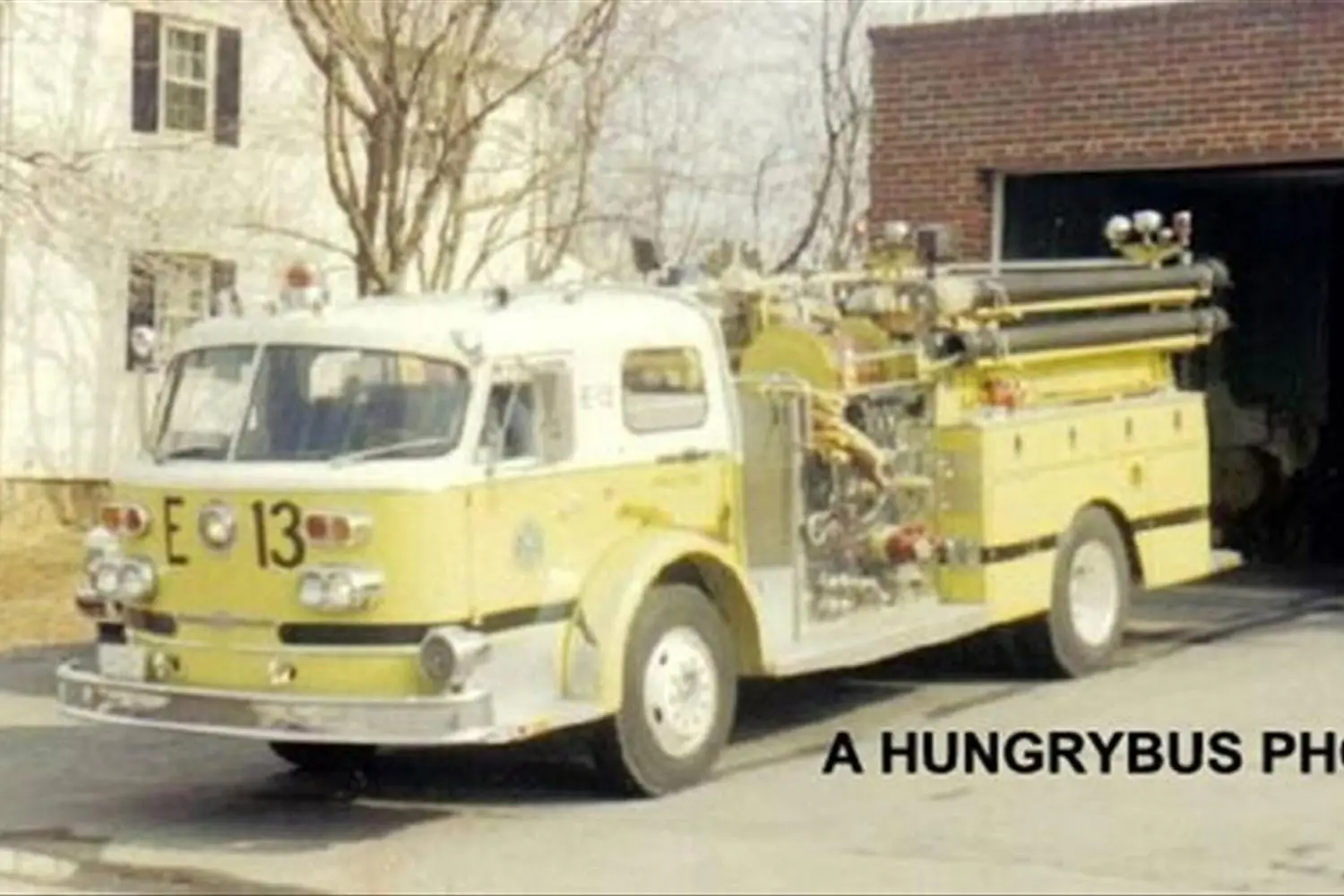 A vintage yellow fire truck with "E 13" printed on the front and sides is parked outside a brick fire station. The truck is equipped with various hoses and equipment. Text reading "A HUNGRYBUS PHOTO" is overlaid on the image.