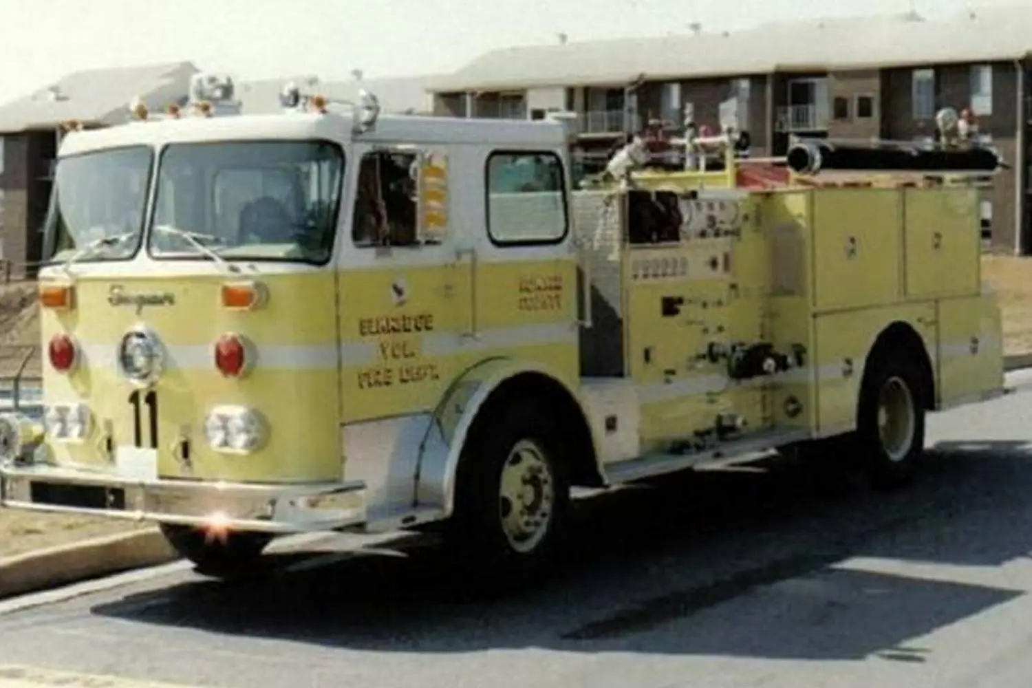 A yellow fire truck with the label "Eldridge Vol. FIRE DEPT." and number "11" on the front. It is parked on a residential street with apartment buildings in the background. The truck has equipment and hoses attached to its side.