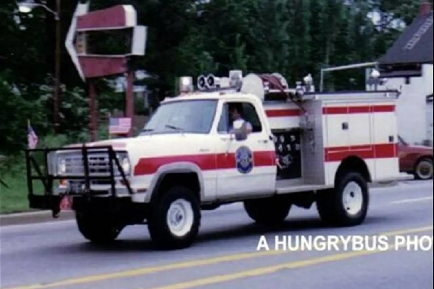 A white and red fire truck is driving down a street, with a person in the driver's seat. The truck has a front bumper guard and firefighting equipment visible on top and sides. Trees and a partially visible sign are in the background. Text on the image reads "A HUNGRYBUS PHOTO.