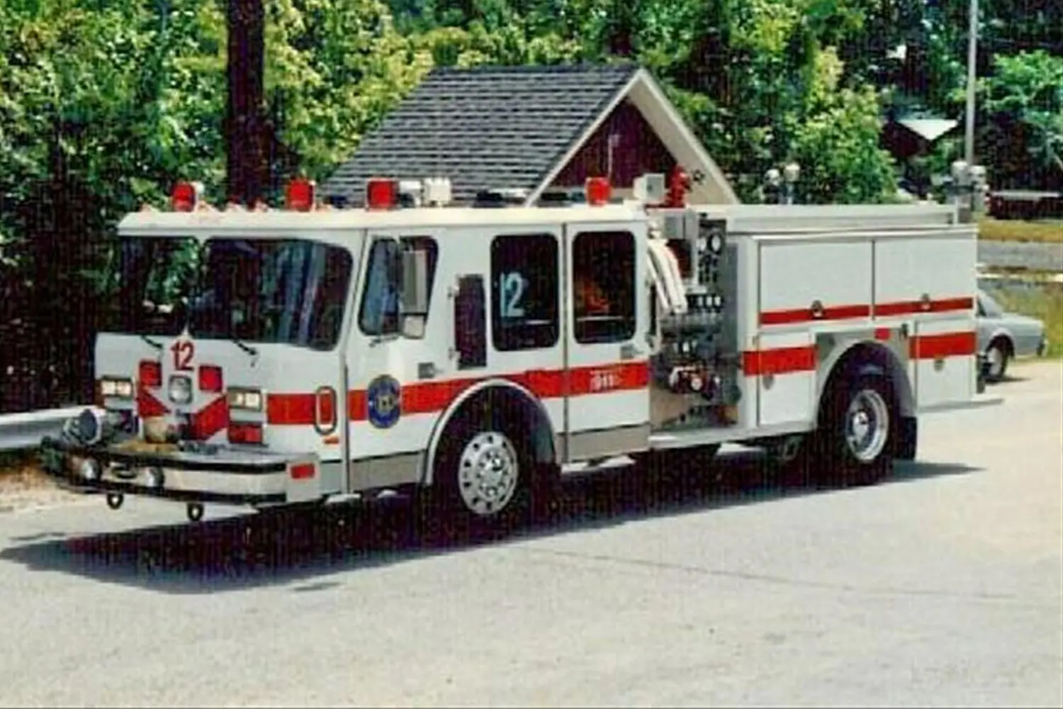 A white fire truck with red stripes and the number 12 on its front, parked on a road. The truck has several red lights on top and various equipment stored on its sides. In the background, there are trees and a small building.