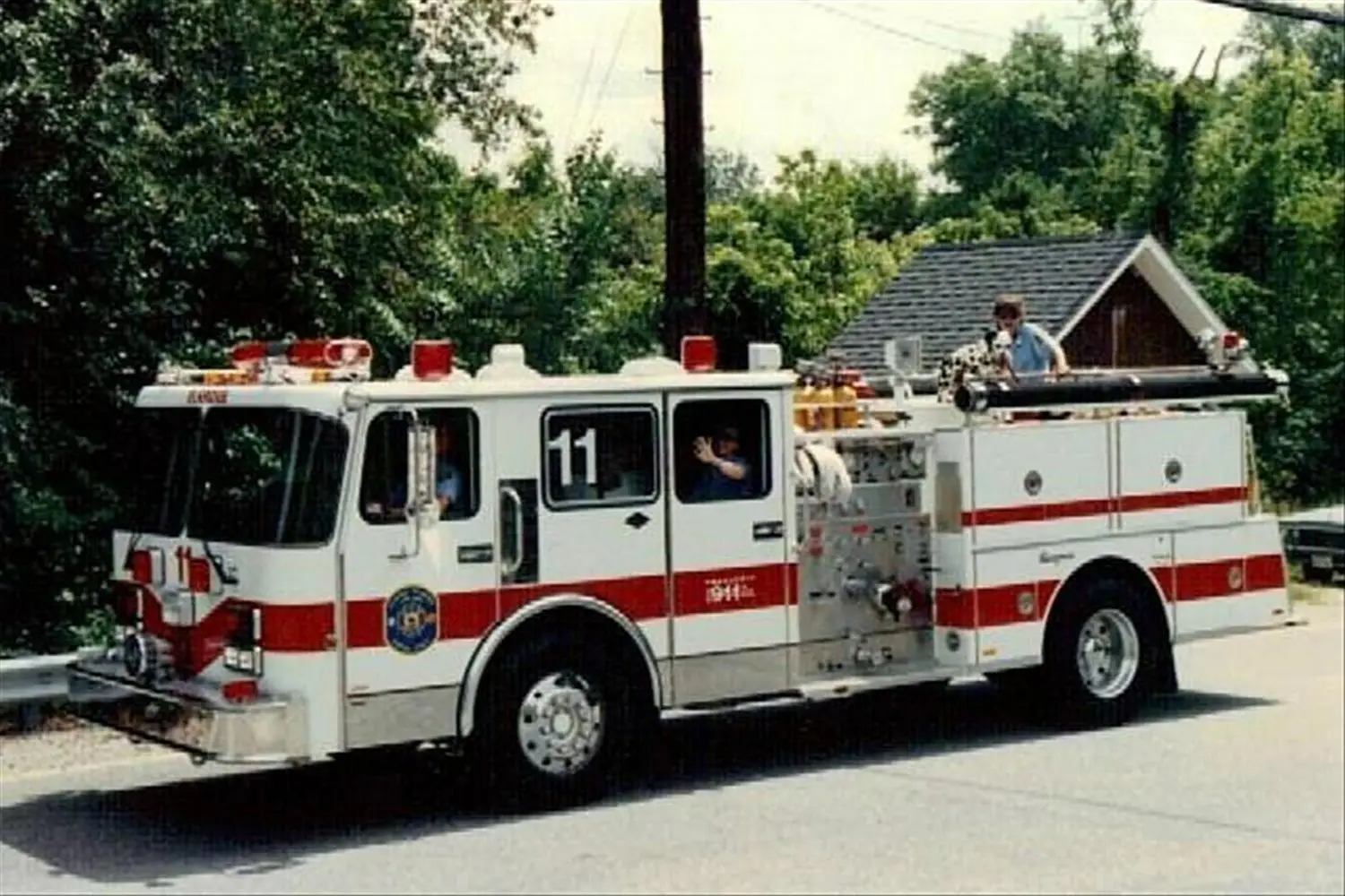 A red and white fire truck labeled "11" is parked on the side of a road. The vehicle features various equipment and hoses. Trees and a small building are visible in the background. Two firefighters can be seen, one seated inside and another standing on the truck.
