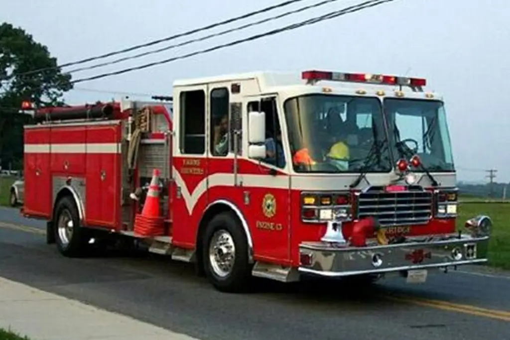 A red and white fire truck is driving down a road. The truck has various firefighting equipment, including hoses and traffic cones. One firefighter is visible in the cab, and emergency lights are mounted on the roof. The truck is marked with "Engine 13.
