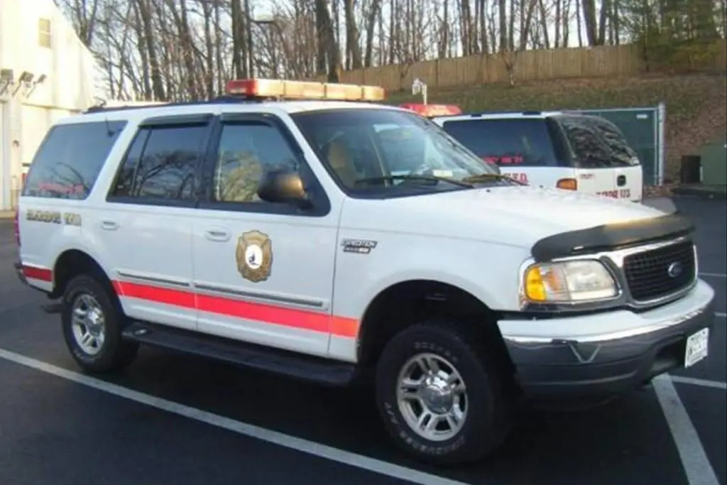 A white Ford SUV with red stripes and emergency lights on the roof is parked in a lot. The vehicle features a badge emblem and the text "Emergency Unit" on the side. Another similar vehicle is partially visible behind it. Trees and a fence are in the background.