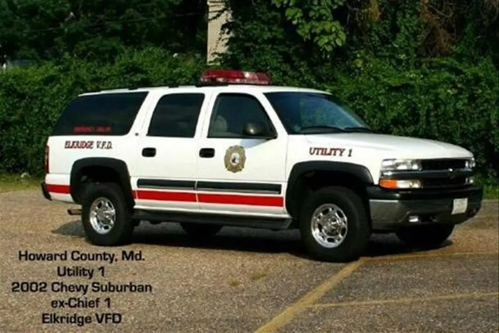 A white SUV with "Elkridge V.F.D" and "Utility 1" markings is parked on a paved area. The vehicle features a red emergency light on top and red stripes on the sides. Text overlay reads "Howard County, Md. Utility 1 2002 Chevy Suburban ex-Chief 1 Elkridge VFD.