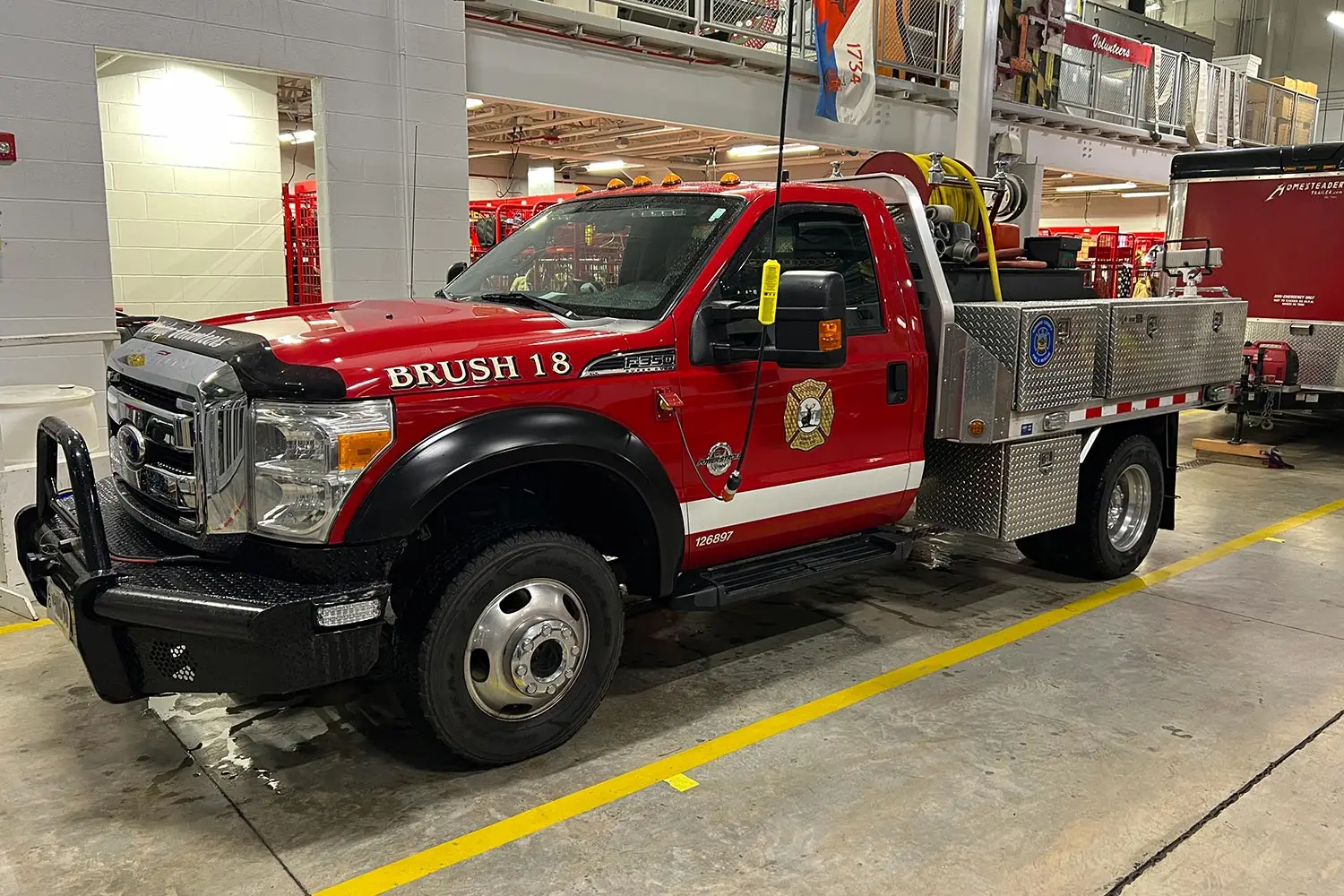 A red and white brush fire truck labeled "Brush 18." The truck is parked indoors and equipped with fire-fighting tools and equipment, including a hose reel, storage compartments, and an emergency light bar. Various firefighting gear is visible in the background.