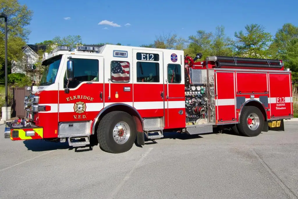 A red and white fire truck from the Elkridge Volunteer Fire Department (E.V.F.D.) is parked on a driveway. The truck is labeled as Engine 12 (E12) and features an American flag, firefighting equipment, and various department insignias. Trees and a house are in the background.