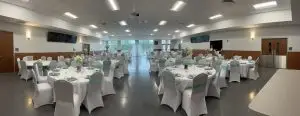 A spacious banquet hall set up for an event, featuring round tables with white tablecloths and teal chair sashes. The tables are elegantly decorated with flowers, candles, and silverware. Large windows at the back provide natural light. The hall is well-lit with ceiling lights.