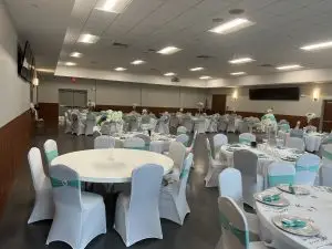 A banquet hall decorated for an event with neatly arranged round tables. Each table is covered with white linen, turquoise-accented centerpieces featuring flowers, and matching place settings. White chairs with turquoise sashes are set up around the tables.