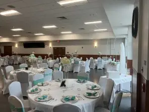A spacious banquet hall set for a formal event. Tables are covered with white cloths, adorned with teal accents, and elegantly arranged with glassware and plates. Floral centerpieces in white and pastel hues decorate the tables. The room has white walls and a tiled floor.