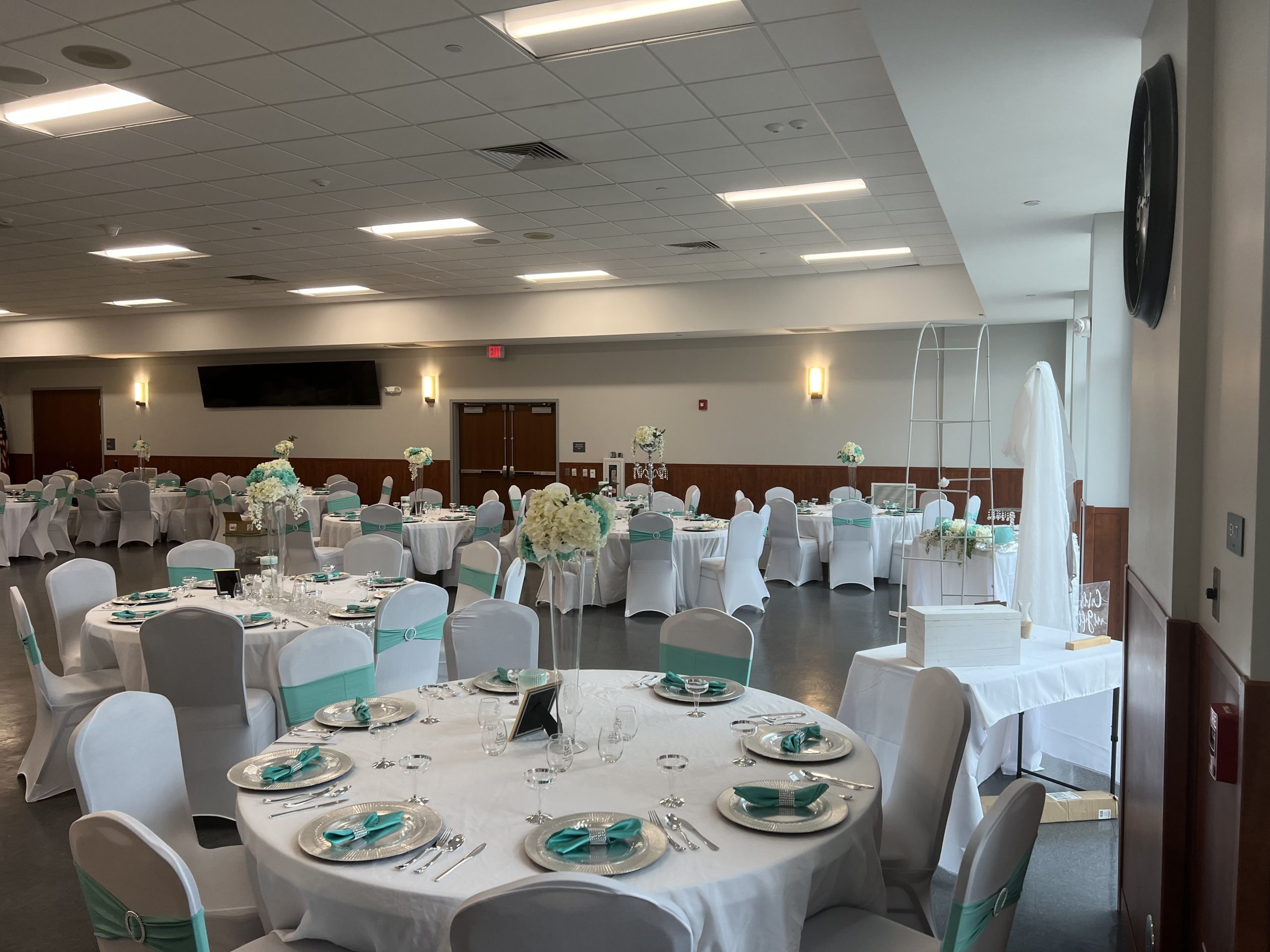 A spacious banquet hall set for a formal event. Tables are covered with white cloths, adorned with teal accents, and elegantly arranged with glassware and plates. Floral centerpieces in white and pastel hues decorate the tables. The room has white walls and a tiled floor.