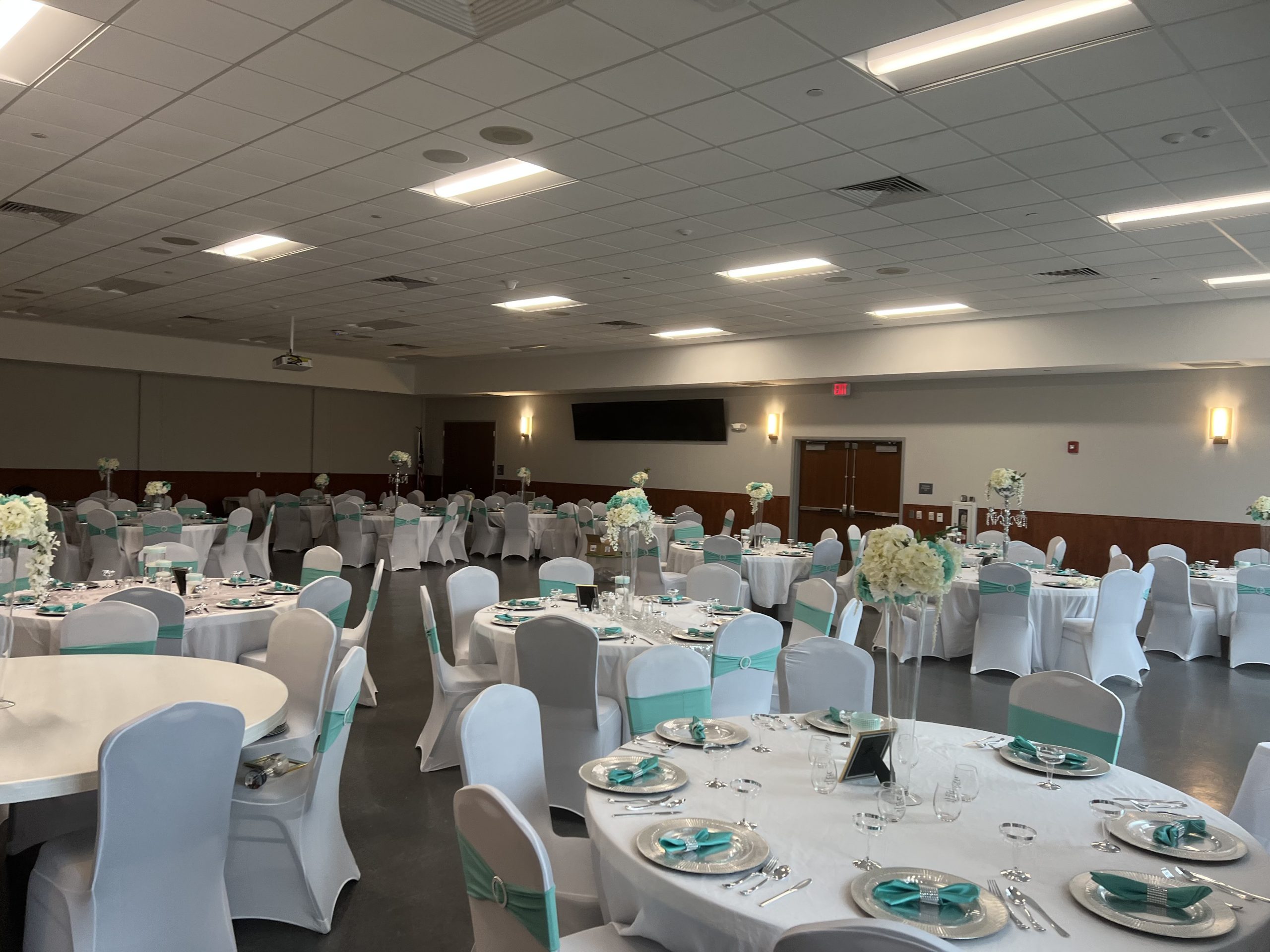 A large event hall decorated for a formal gathering with round tables and white chairs. Tables are adorned with white tablecloths, turquoise napkins, and white floral centerpieces. Ceiling lights illuminate the space, creating a bright and elegant atmosphere.