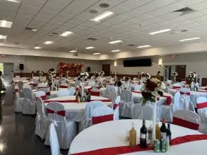 A spacious banquet hall with round tables covered in white tablecloths and red sashes. Each table has floral centerpieces and bottles of beverages. A decorated backdrop with floral arrangements and balloons is visible at the front of the room.