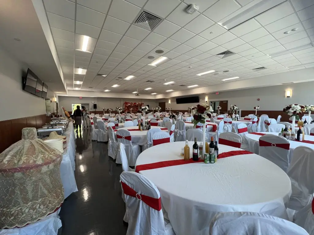 A spacious banquet hall set up for an event. Round tables draped in white tablecloths and adorned with red sashes are arranged throughout. Tables feature centerpieces and bottles. A buffet area is visible on the left, and the room is illuminated by overhead lighting.