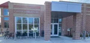 Exterior view of Elkridge Community Hall. The entrance features a glass door and windows, with a brick façade and a metal canopy. Patio tables and chairs are situated to the left of the door, and a bike rack is visible on the right.