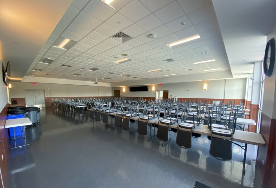 A spacious dining hall with rows of long tables and chairs stacked upside down on each table. The room has a modern design with a tiled ceiling, large windows on the right, and a polished floor. The walls have a mix of white and wooden panels.