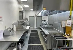 A clean and organized commercial kitchen with stainless steel countertops and appliances. There are cooking stations with a stove, grill, and prep areas on both sides. Rubber mats line the floor, and a couple of sinks are visible along the left side wall.