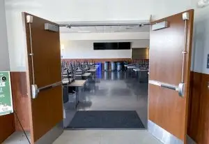 A double-door entrance leads into an empty dining area. Tables and chairs are stacked, with chairs placed upside down on the tables. Several large trash bins line the back wall. Walls are neutral-colored, and the floors are polished.