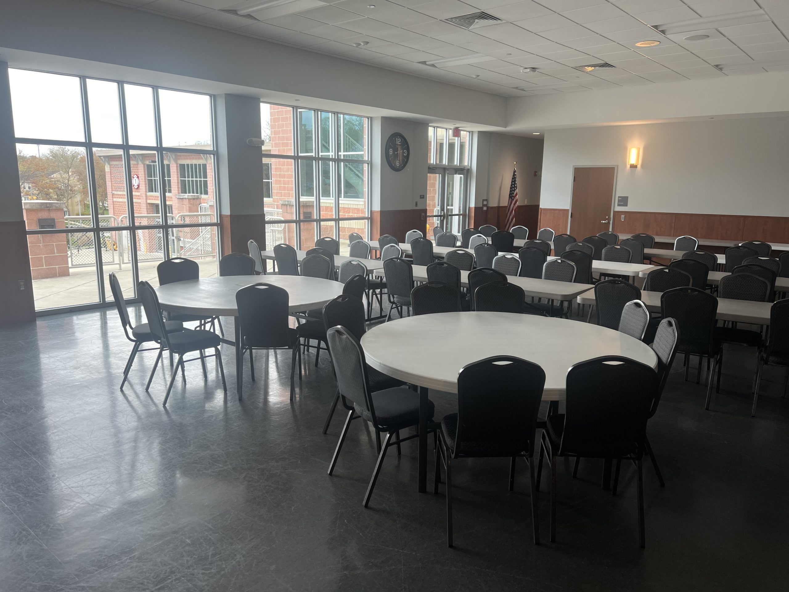A spacious room with large windows, round tables, and black chairs arranged in rows. There is a flag in the corner, and the room is well-lit, with a view of the outdoors through the windows. The floor is tiled, and lighting fixtures are mounted on the ceiling.