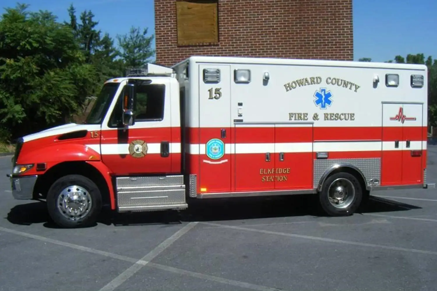 A fire and rescue truck from Howard County is parked on the street. The vehicle is white with a red stripe and has "Howard County Fire & Rescue" written on the side. The truck features emergency medical marks and the number 15. Trees and a brick building are in the background.