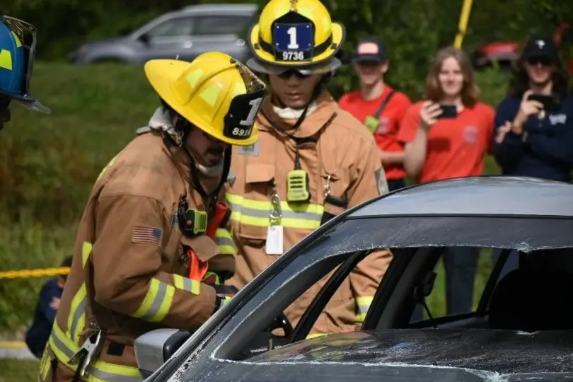 Firefighters in yellow helmets and brown uniforms assess a damaged car at an outdoor scene. One firefighter, holding a tool, works near the car's window, while the other observes. Bystanders in the background take photos and watch.