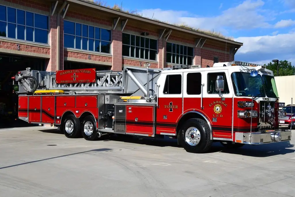 A large red fire truck with extended ladder parked in front of a brick fire station. The truck has multiple compartments and emergency lights on the roof. "Walker Fire Department" is inscribed on the side. The fire station has multiple garage doors.