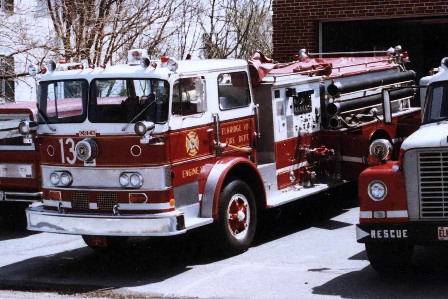 A vintage fire engine labeled "Engine 13" with a white and red color scheme is parked in front of a brick building. The vehicle has numerous hoses and equipment on its sides. Nearby, a smaller red rescue vehicle is partially visible.