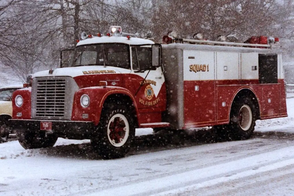 A red and white fire truck marked "Squad 1" and "Randolph Vol. Fire Dept." is parked on a snow-covered street with trees and parked cars in the background. Snow is actively falling, covering the ground and parts of the truck.