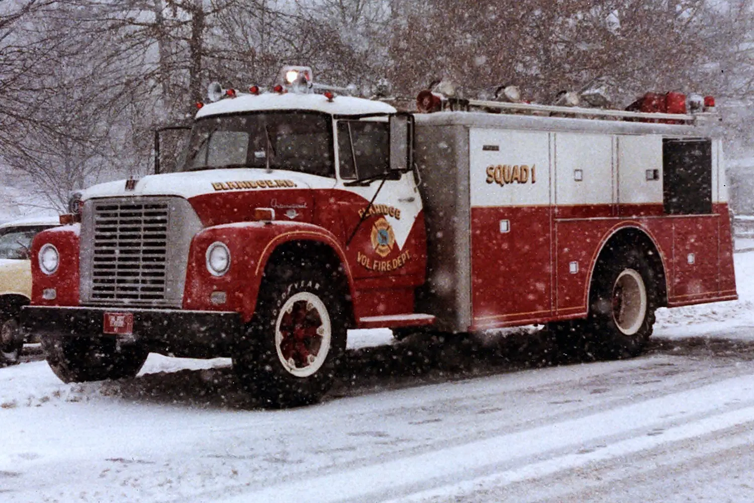 A red and white fire truck marked "Squad 1" and "Randolph Vol. Fire Dept." is parked on a snow-covered street with trees and parked cars in the background. Snow is actively falling, covering the ground and parts of the truck.