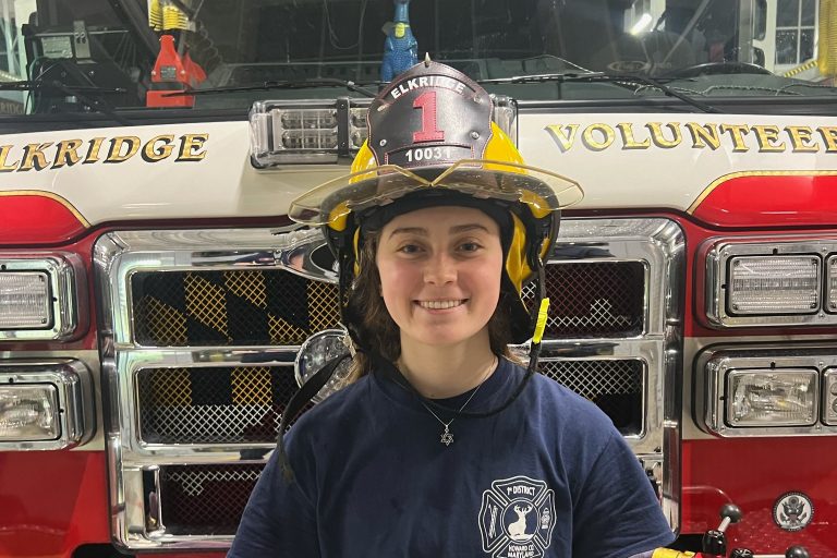 A firefighter in uniform stands in front of a fire truck. The individual is wearing a helmet labeled "ELKRIDGE VOLUNTEER." The fire truck has visible equipment and hoses, with the words "ELKRIDGE VOLUNTEER" partially visible on the front.