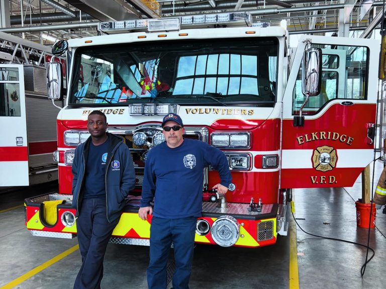 Two firefighters wearing dark uniforms stand in front of a red and white fire truck inside a fire station. The truck has "Elkridge Volunteers" and "Elkridge V.F.D." written on it. The station has shiny floors, high ceilings, and various gear and equipment.