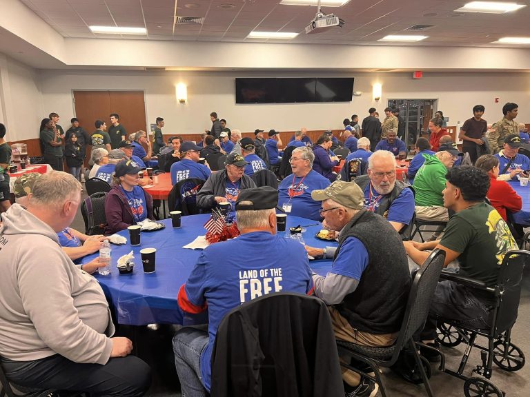 A large group of people, mainly older adults, are seated at round tables in a community hall. They are engaged in conversation and enjoying meals. Many are wearing blue shirts with "Land of the Free" printed on them. There are also people standing and interacting in the background.