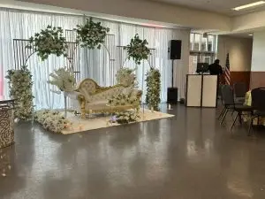 A decorated wedding stage features a white and gold ornate loveseat surrounded by white floral arrangements and greenery. Candles are placed around the stage, and a DJ setup with speakers is visible in the background. Rows of chairs are arranged in front.