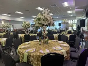 A banquet hall adorned with elegantly set tables covered in gold tablecloths, each featuring floral centerpieces with white roses. Champagne bottles and glasses are on the tables, and the chairs are neatly arranged around them. The room is ready for an event.
