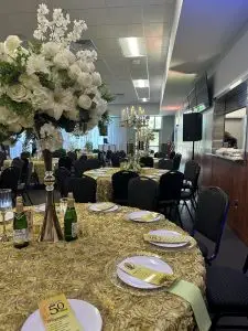 A decorated banquet hall with round tables covered in gold textured tablecloths is set up for a formal event. Each table features large floral centerpieces, champagne bottles, and place settings with menus. Black chairs surround the tables.