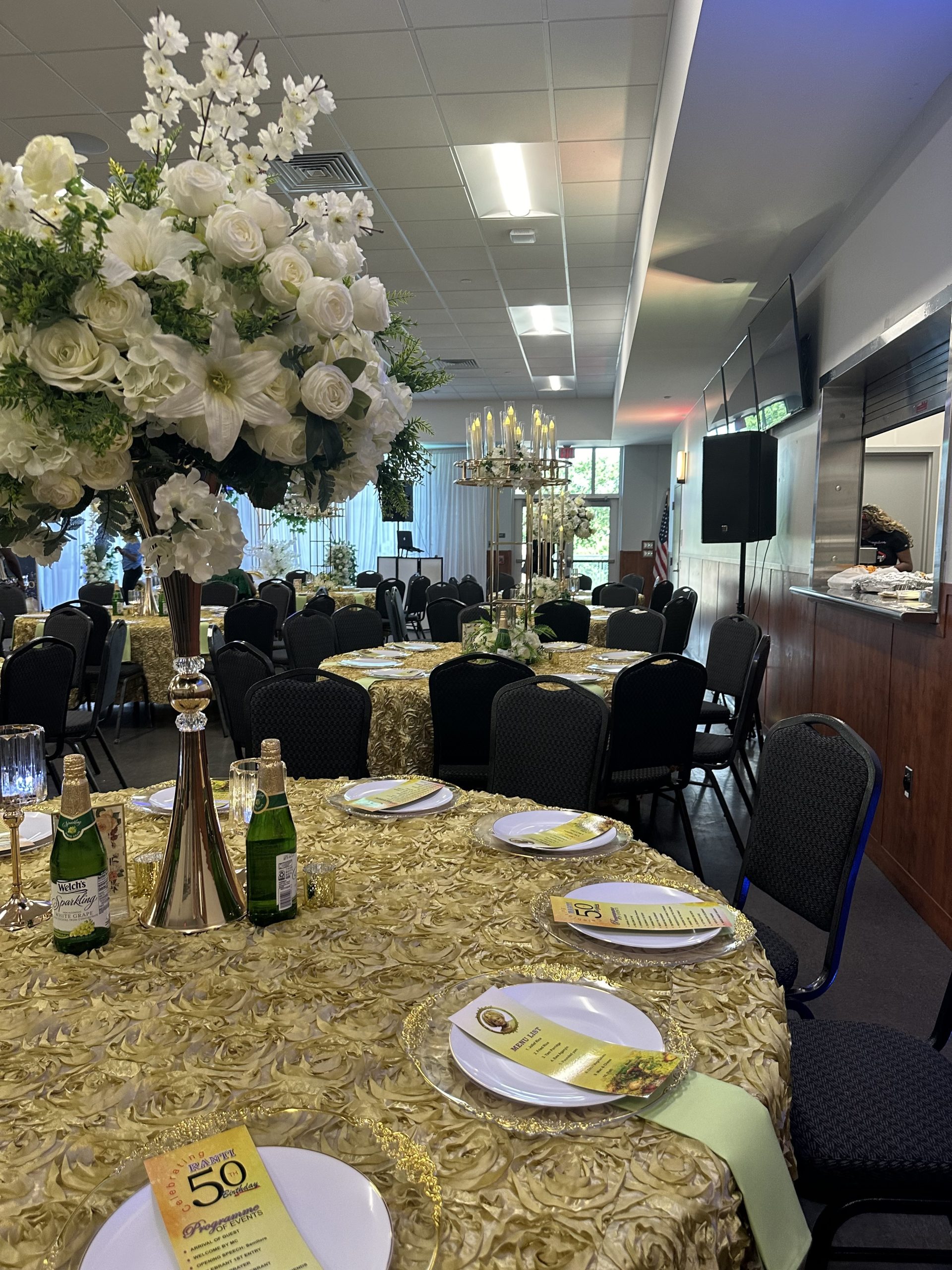 A decorated banquet hall with round tables covered in gold textured tablecloths is set up for a formal event. Each table features large floral centerpieces, champagne bottles, and place settings with menus. Black chairs surround the tables.