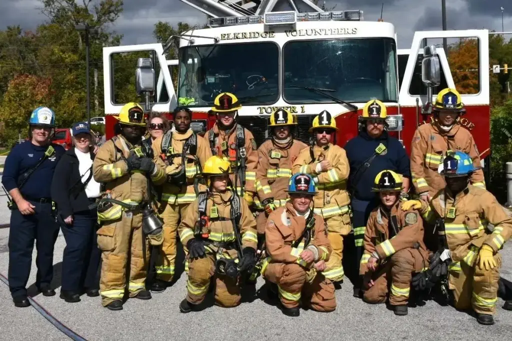 A group of firefighters, both men and women, pose in front of a fire truck labeled "BERKIDGE VOLUNTEERS." The firefighters are wearing various gear, including helmets, fire jackets, and gloves, and appear to be smiling for the photograph. Trees can be seen in the background.