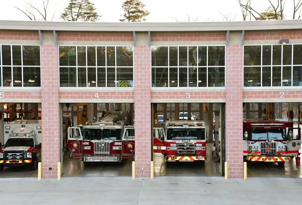A fire station with four fire trucks parked inside four glass-fronted bays, numbered 2 to 5 from right to left. The brick building features large windows above the bays, and some trees are visible in the background.