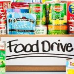 A cardboard box labeled "Food Drive" is filled with various canned and packaged foods, including vegetables, soup, sardines, fruit, and crackers, with more cans placed around the box.