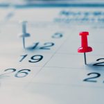 Close-up of a calendar page with push pins marking specific dates. A red push pin is on the 20th, while white pins mark the 12th and 26th. The focus is sharp on the pins and dates.